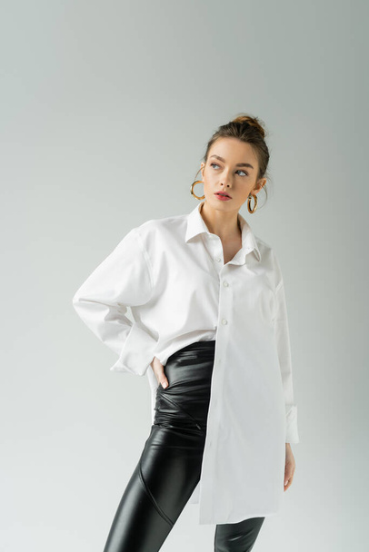 trendy woman in white shirt and black pants posing with hand on hip isolated on grey - Fotó, kép