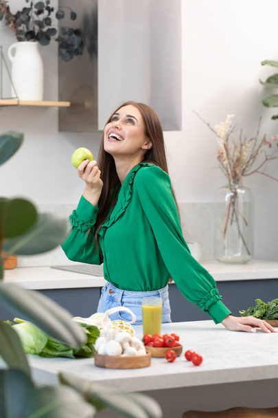 A happy young woman holds a green apple in her hand in the kitchen. The concept of healthy eating, vegetarianism. - Foto, imagen