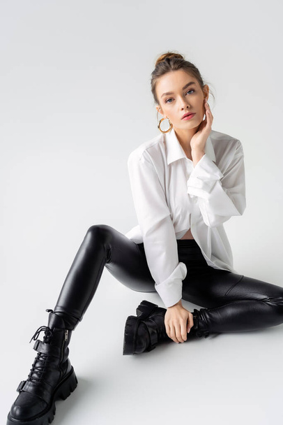 full length of young woman in white shirt and black latex pants with rough boots sitting on grey background - Fotó, kép