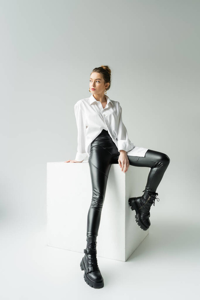 pełna długość modelu w białej koszuli oversize i czarne spodnie z szorstkimi butami siedzącymi na białym sześcianie na szarym tle - Zdjęcie, obraz