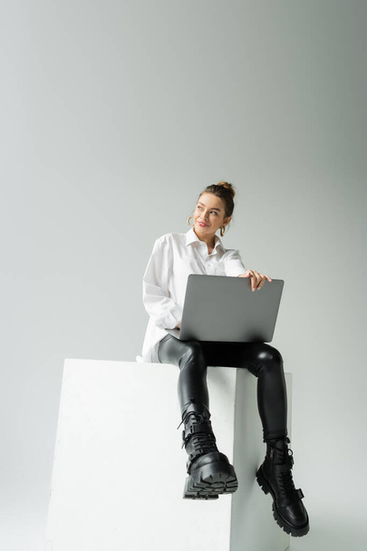 longitud completa de la mujer feliz en pantalones de látex negro y botas de cuero sentado en cubo blanco con portátil sobre fondo gris - Foto, imagen