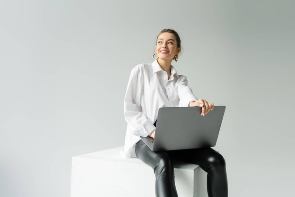 cheerful woman in trendy outfit sitting on white cube with laptop and looking away isolated on grey - Foto, Imagem