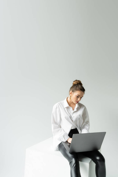 Geniş tişörtlü ve dar pantolonlu genç bir kadın gri arka planda küp üzerinde otururken dizüstü bilgisayarla çalışıyor. - Fotoğraf, Görsel