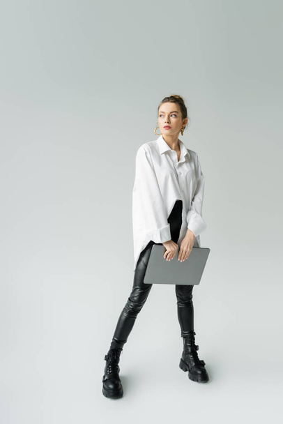 pełna długość kobiety w białej koszuli oversize i czarne skórzane buty stojące z laptopem na szarym tle - Zdjęcie, obraz
