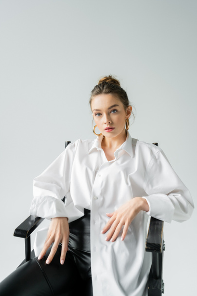 stylish woman in white shirt and golden earrings sitting on chair and looking at camera isolated on grey - Fotoğraf, Görsel