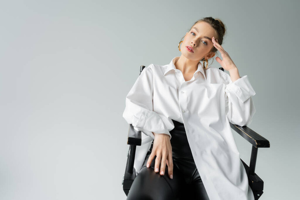 pretty woman in white oversize shirt and black tight pants posing on chair with hand near head isolated on grey - Fotografie, Obrázek