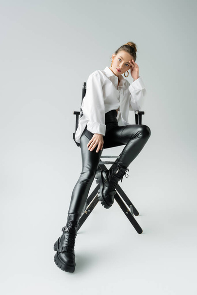 full length of stylish woman in white shirt and black pants with leather boots sitting on chair on grey background - Photo, Image
