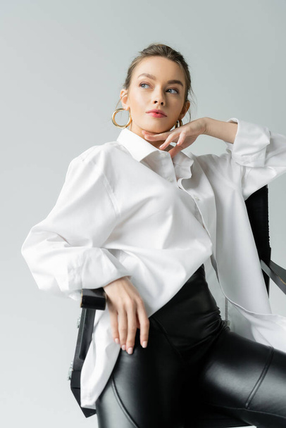 young woman in oversize shirt touching chin and looking away while sitting on chair isolated on grey - Foto, imagen