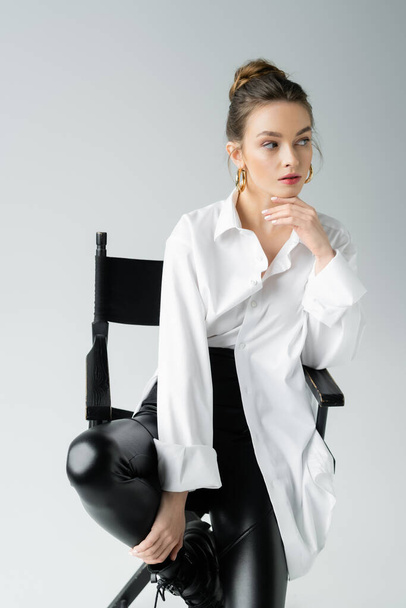 mujer de moda en camisa blanca y pantalones de látex negro sentado en la silla y mirando hacia otro lado aislado en gris - Foto, imagen