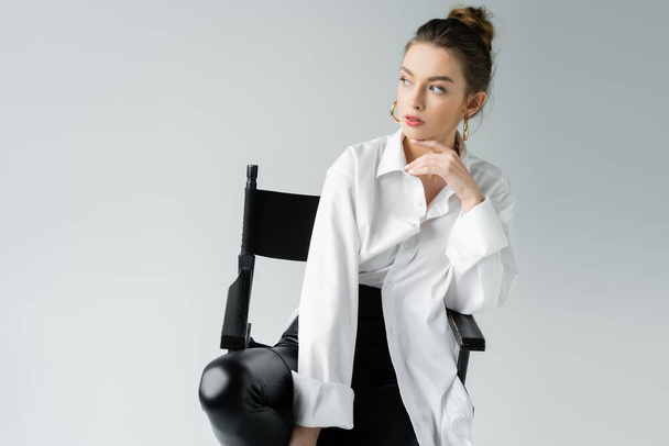 młoda kobieta w koszuli oversize i czarne obcisłe spodnie trzymając się za ręce w pobliżu podbródka siedząc na krześle odizolowana na szaro - Zdjęcie, obraz