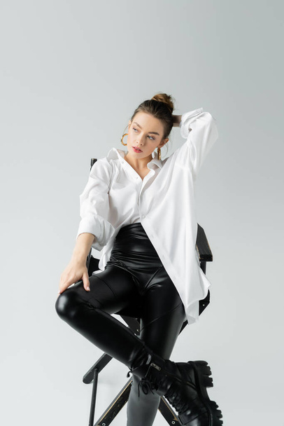 stylish woman in black tight pants and white shirt holding hand behind head while sitting on chair on grey background - Fotó, kép