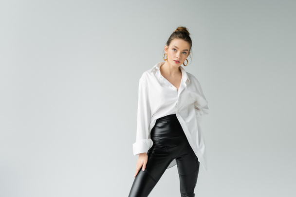 stylish woman in black tight pants and white oversize shirt posing and looking at camera isolated on grey - Foto, Imagem