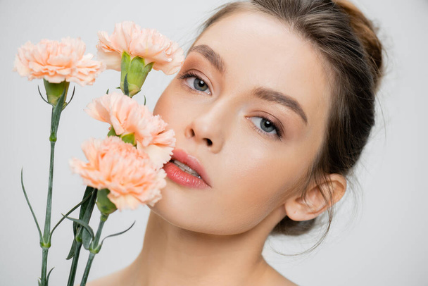 young woman with perfect skin and natural makeup looking at camera near fresh carnations isolated on grey - Fotoğraf, Görsel