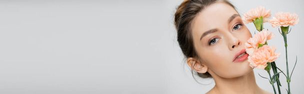 молодая и красивая женщина с натуральным макияжем, смотрящая в камеру рядом со свежими гвоздиками, изолированными на сером, баннер - Фото, изображение