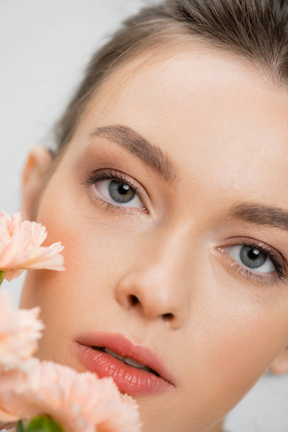 close up portrait of young woman with natural makeup looking at camera near blurred flowers isolated on grey - Fotoğraf, Görsel
