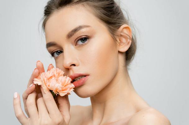 mujer sensual con piel perfecta y maquillaje natural sosteniendo clavel fresco y mirando a la cámara aislada en gris - Foto, imagen