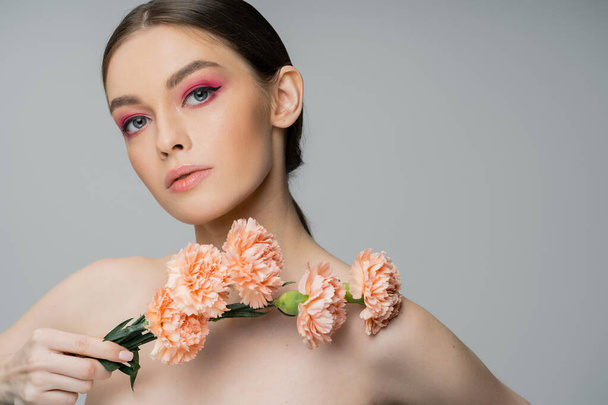 молодая женщина с макияжем и обнаженными плечами позирует со свежими цветами и смотрит на камеру, изолированную на сером - Фото, изображение