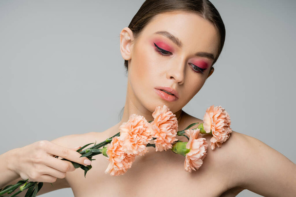 pretty woman with pink visage holding fresh carnations near bare shoulder isolated on grey - Foto, Bild