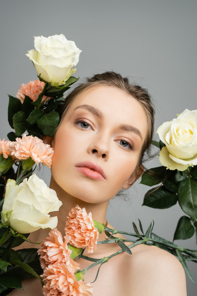 young woman with natural makeup looking at camera near fresh flowers isolated on grey - Photo, Image