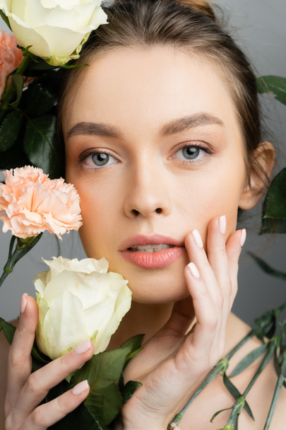 portrait of pretty woman with perfect skin touching face near roses and carnations isolated on grey - Photo, Image