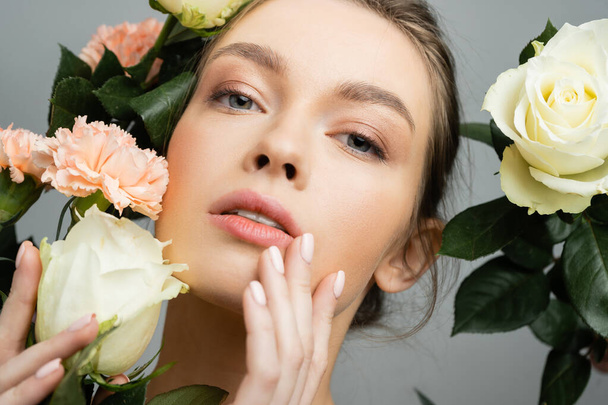 portrait of sensual woman touching face while looking at camera near roses and carnations isolated on grey - Foto, afbeelding