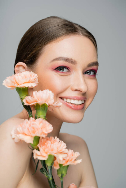 happy young woman with makeup and perfect skin smiling at camera near peach carnations isolated on grey - Photo, Image
