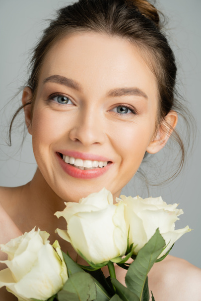 Πορτρέτο της χαρούμενης νεαρής γυναίκας κοιτάζοντας κάμερα κοντά τριαντάφυλλο λουλούδια απομονώνονται σε γκρι  - Φωτογραφία, εικόνα