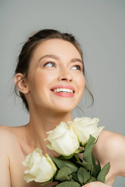 Femme heureuse aux épaules nues tenant des roses et regardant loin isolé sur gris  - Photo, image