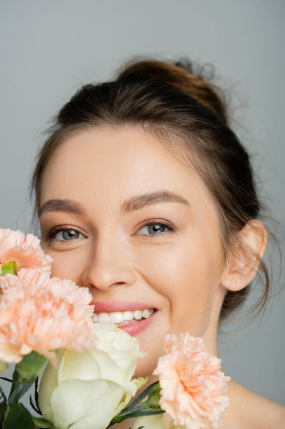 Cheerful young woman looking at camera near blurred bouquet isolated on grey  - Valokuva, kuva