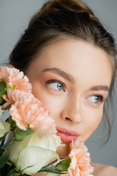 Retrato de una joven bonita mirando hacia otro lado cerca de flores aisladas en gris  - Foto, Imagen