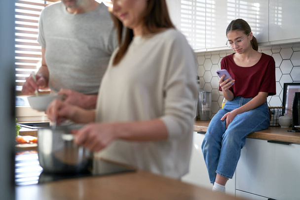 Adolescente utilisant le téléphone portable dans la cuisine lorsque les parents cuisinent ensemble  - Photo, image