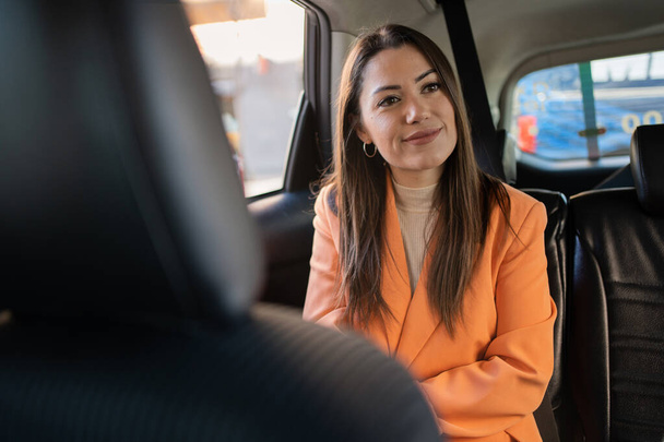 woman inside the back of a car, transport by cab or car with driver, young girl talking to cab driver animatedly and smiling. Person traveling for work, dressed in orange blazer and cream sweater. Very pretty and natural girl. - Photo, Image
