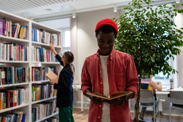 Iloinen Afrikkalainen Amerikkalainen mies seisoo yliopiston kampuksella lukea saada uusia taitoja, parantaa vieraiden kielten tuntemusta taustalla kirjastonhoitaja järjestää kirjoja oikein hyllyillä lomakkeet tilauksia opiskelijoille - Valokuva, kuva