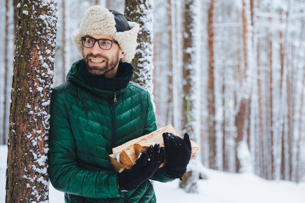 Bearded young male wears warm hat and green jacket, holds pile of firewood, looks happily aside, notices something, likes winter and frosty weather. Dreamful attractive man spends time in forest - Photo, Image