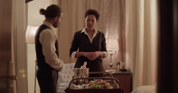Elegant young African American female guest talking to friendly waiter standing near trolley with wine and fruits during room service at hotel - Video