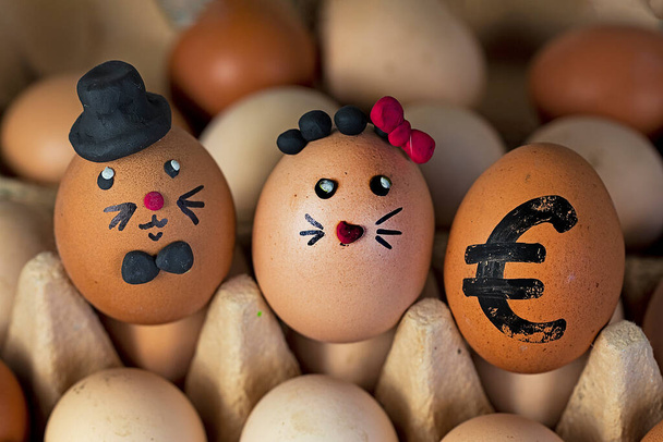 δύο χαριτωμένα αυγά λαγού σε ένα μαύρο καπέλο και με ένα κόκκινο τόξο πλαστελίνης σε ένα δοχείο αυγών με ένα ζωγραφισμένο αυγό ευρώ. Αύξηση στην τιμή. Ίστα - Φωτογραφία, εικόνα