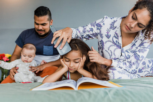 spanyol család olvas egy könyvet, miközben anya fésülködik a lánya haját az ágyon otthon Mexikóban Latin-Amerikában - Fotó, kép