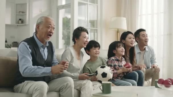 three generation asian family sitting on couch at home watching live broadcasting of soccer game together celebrating a goal - Video