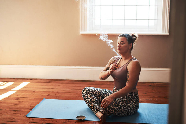 Palo santo benefits include detoxification and reducing stress. a young woman burning a palo santo stick while sitting on a yoga mat at home - Foto, imagen
