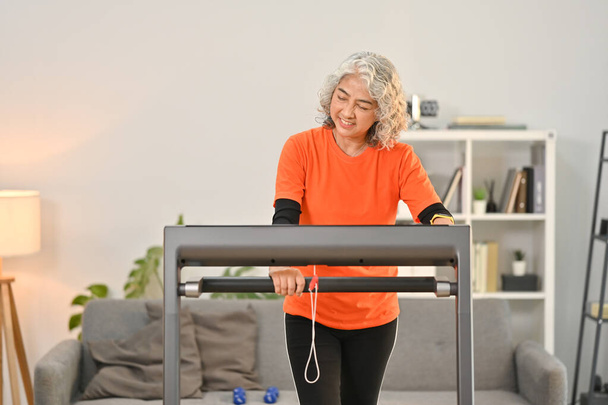 Smiling middle aged woman in sport clothes walking on treadmills in the morning at home. Healthy lifestyle concept. - Foto, imagen