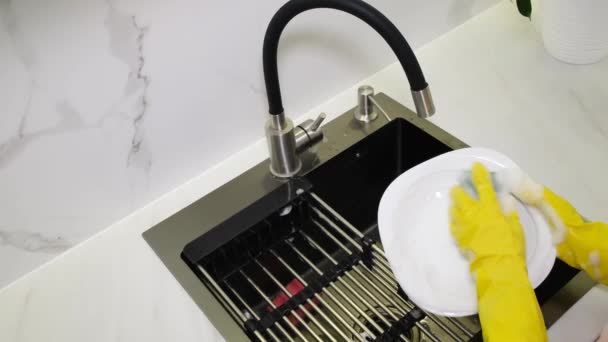 Κοντινό πλάνο γυναικείων χεριών που πλένουν βρώμικα πιάτα στο νεροχύτη της κουζίνας. Τα γυναικεία χέρια πλένουν ένα πιάτο κάτω από τη βρύση. Υπηρεσίες καθαρισμού κατοικιών - Πλάνα, βίντεο