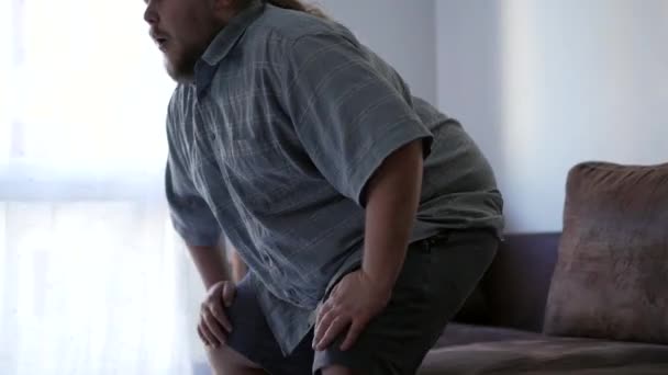 Hombre con dolor de espalda. Una persona con sobrepeso que se levanta del sofá y sufre de lesiones físicas en el interior de su casa. Estilo de vida sedentario - Metraje, vídeo