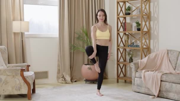 Mujer hermosa y deportiva vestida con polainas negras y un top amarillo, haciendo yoga en la sala de estar. La joven madre entrena individualmente en casa, manteniendo su equilibrio en una pierna. - Metraje, vídeo