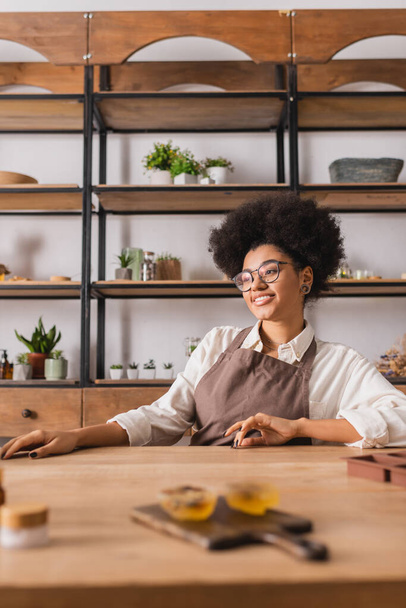 joyful african american craftswoman in apron and eyeglasses sitting near blurred handmade soap on table in workshop - Fotó, kép