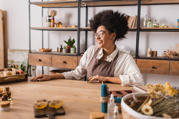 счастливая африканская американская ремесленница, сидящая в мастерской рядом с мылом ручной работы и натуральными ингредиентами на размытом переднем плане - Фото, изображение