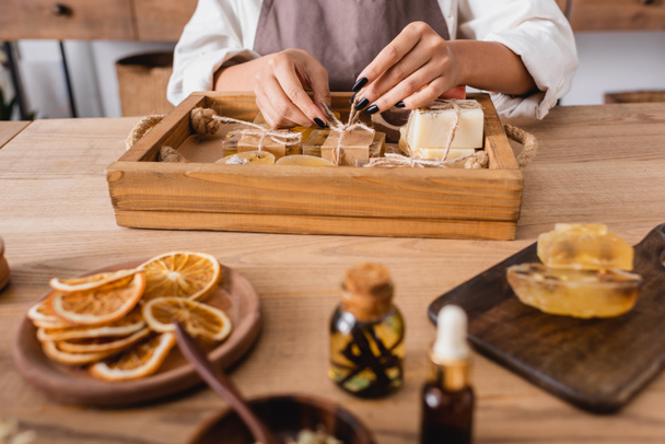 cropped view of african american craftswoman tying twine on handmade soap near dried orange slices and essential oils on blurred foreground - Foto, Imagem