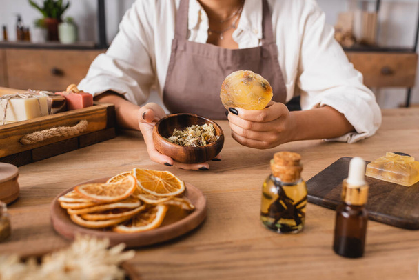partial view of african american woman holding homemade soap and wooden bowl with dried herbs near dried orange slices and essential oils - Foto, imagen