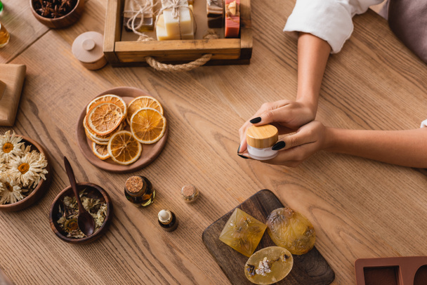 high angle view of cropped african american woman holding cosmetic cream near handmade soap and natural ingredients on wooden table - Foto, Imagem