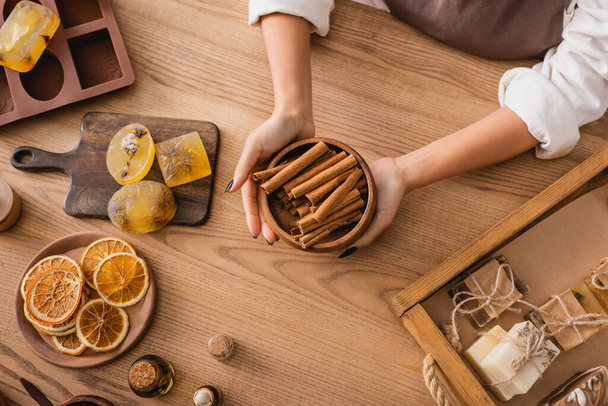 top view of cropped african american woman holding cinnamon sticks near dried orange slices and handmade soap on wooden table - Foto, Imagen