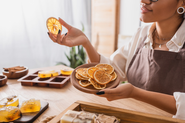 cropped view of smiling african american woman holding wooden plate with dried orange slices near handmade soap in workshop - Foto, afbeelding